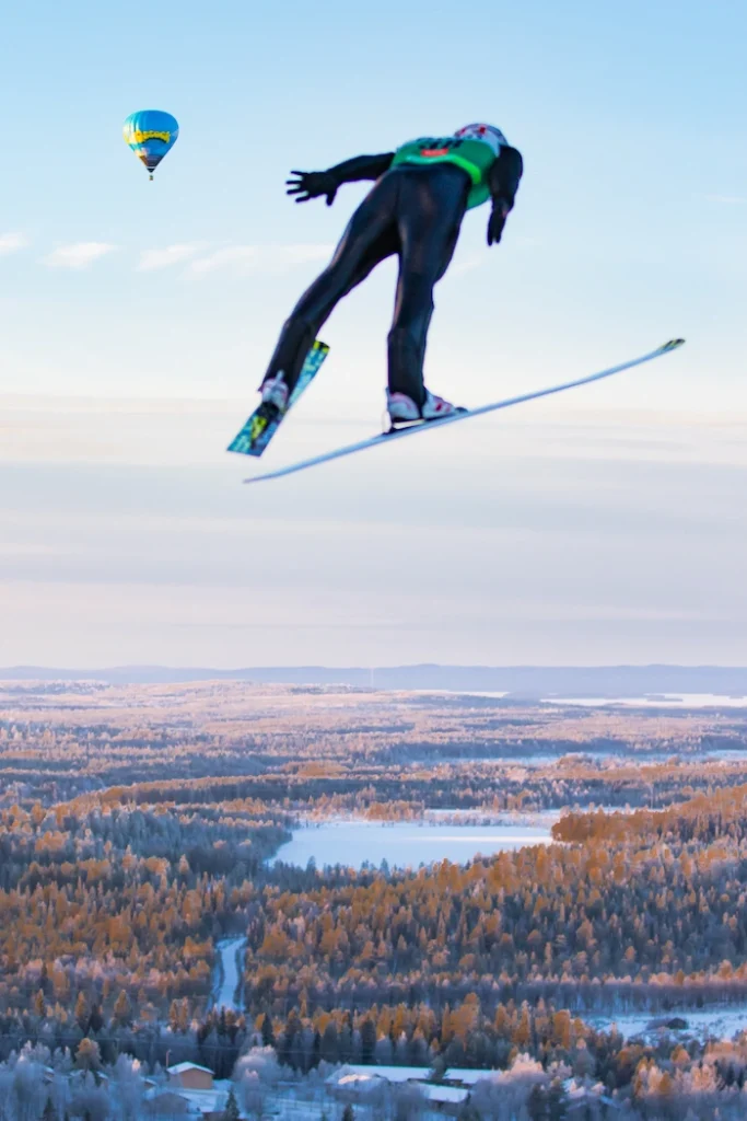 Mäkihyppääjä liitää korkealla taivaalla ja taustalla näkyy talvista maisemaa sekä kuumailmapallo taivaalla.