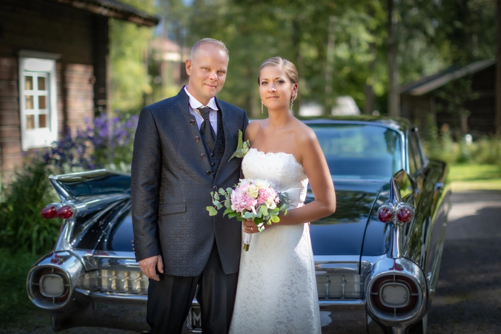 Hääpari seisoo vanhan jenkkiauton edessä ja taustalla näkyy vanhoja rakennuksia sekä metsää.