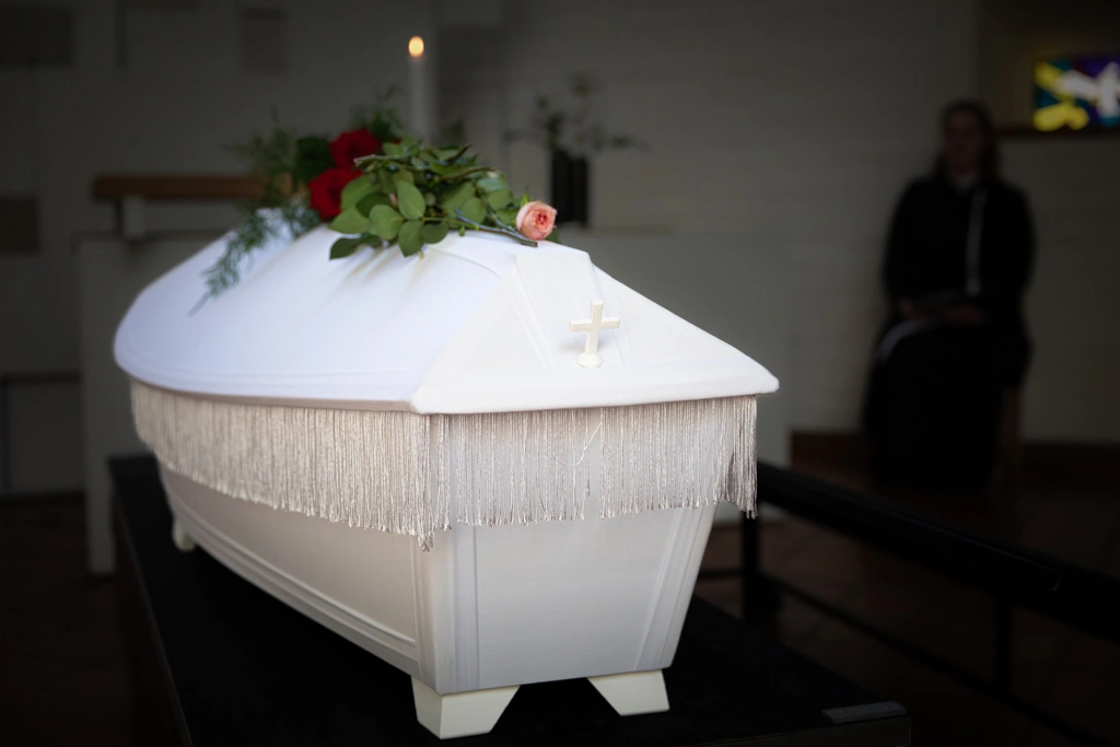 Valkoinen hautausarkku, jonka päällä on kukkia ja taustalla näkyy yksi kynttilä.