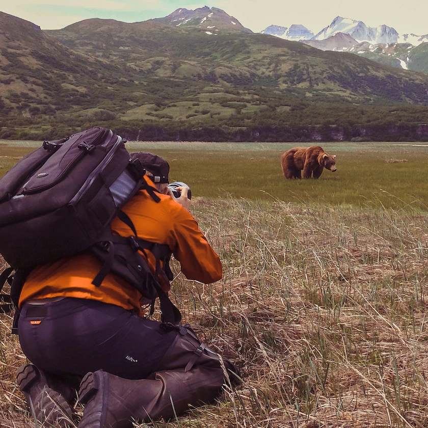 Mies on valokuvaamassa polviltaan karhua, joka on lähellä edessä ja taustalla näkyy vuoristomaisema.