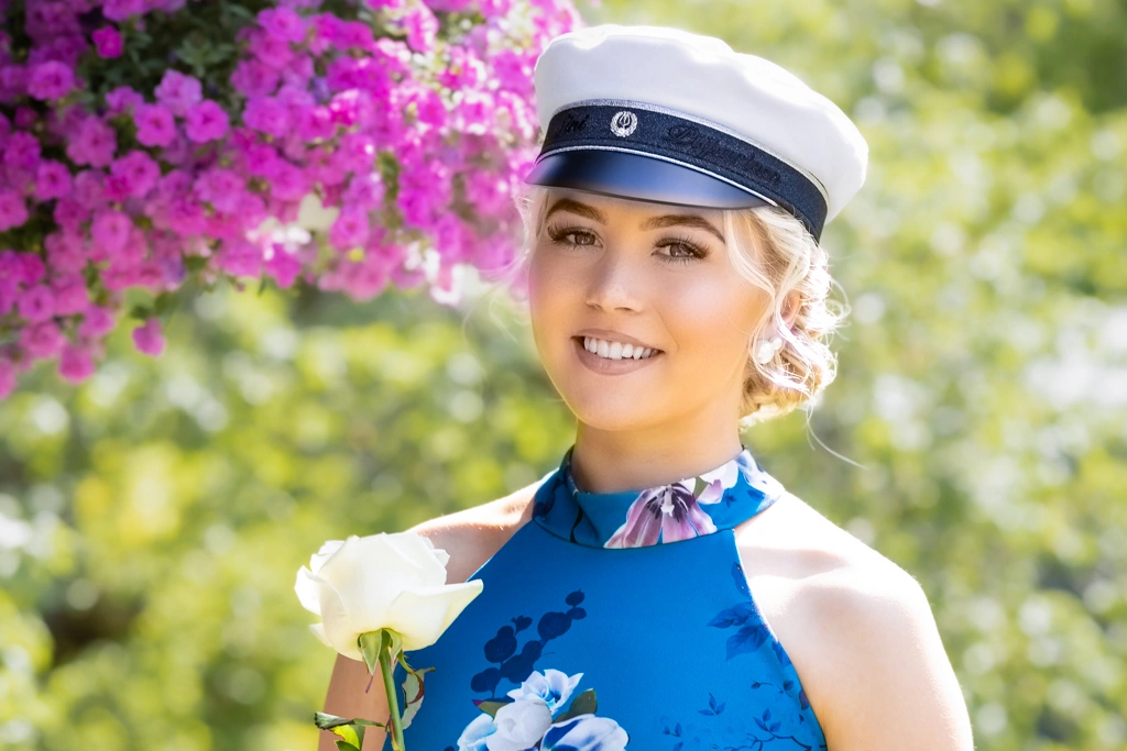 Nainen jolla on ylioppilaslakki päässään ja taustalla näkyy violetti kukka-asetelma sekä vihreitä puiden lehtiä.  