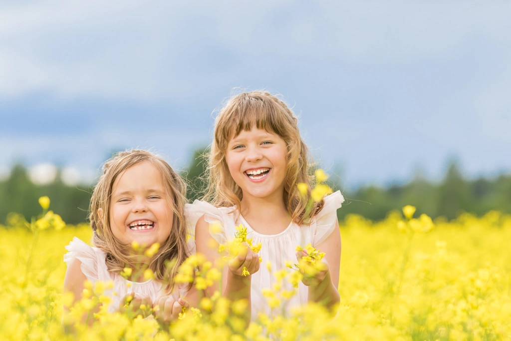 Kaksi nauravaa lasta keltaisen rypsikukkapellon keskellä vaaleissa mekoissaan.