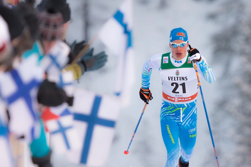 Kuvan vasemmassa laidassa on katsojia ja Suomenlippuja ja oikeassa laidassa on hiihtäjä Iivo Niskanen hiihtämäss numerolappu 21 yllään.