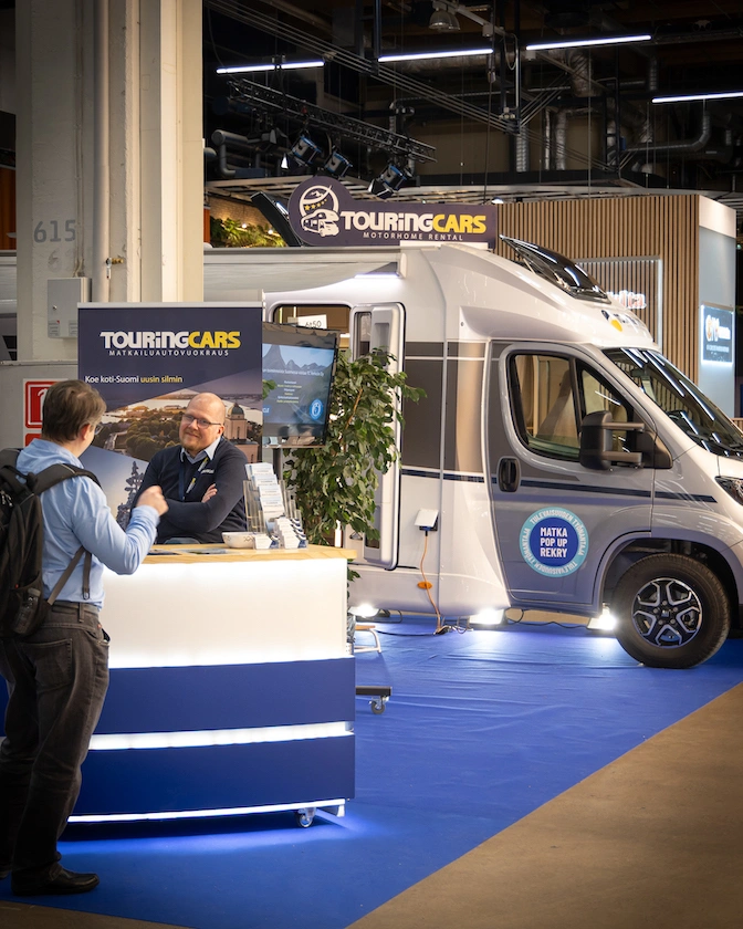 Kaksi miestä juttelevat palvelutiskin äärellä ja taustalla näkyy osittain matkailuauto.