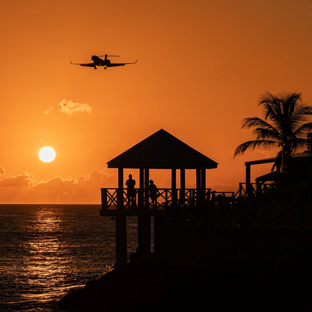 Auringonlasku, jossa näkyy meri, lentokone ja huvimaja siluettina.