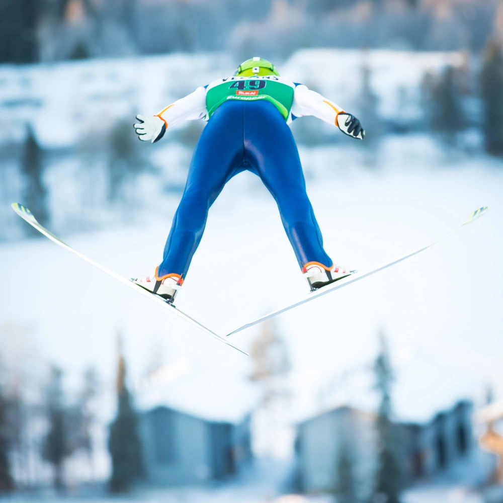 Mäkihyppääjä kuvattuna takanta päin lentäämässä V-asennossa.