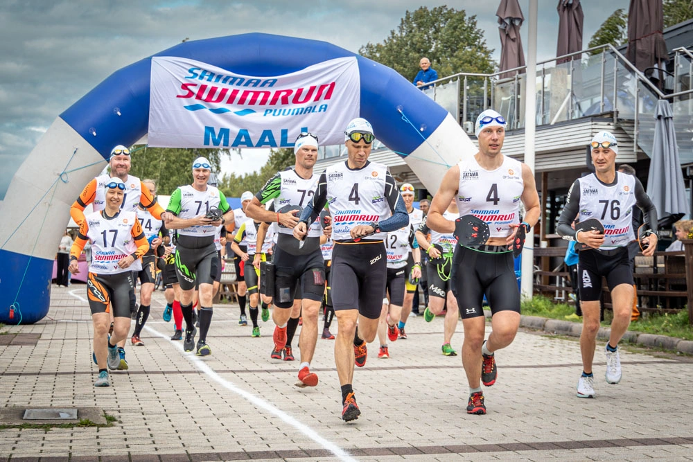 Juoksemassa usaen juoksijan porukka, joilla on uimalakit päässä ja taustalla näkyy Saimaa SwimRun-tapahtuman maalikaari.